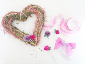 this image shows the supplies needed to make a DIY heart-shaped pink and violet spring wreath with paper roses and a ribbon bow